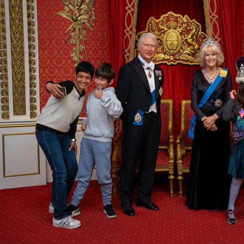 Människor som poserar med kungliga vaxfigurer i ett utsmyckat rum; en person tar en selfie medan en annan placerar en krona på ett barns huvud.