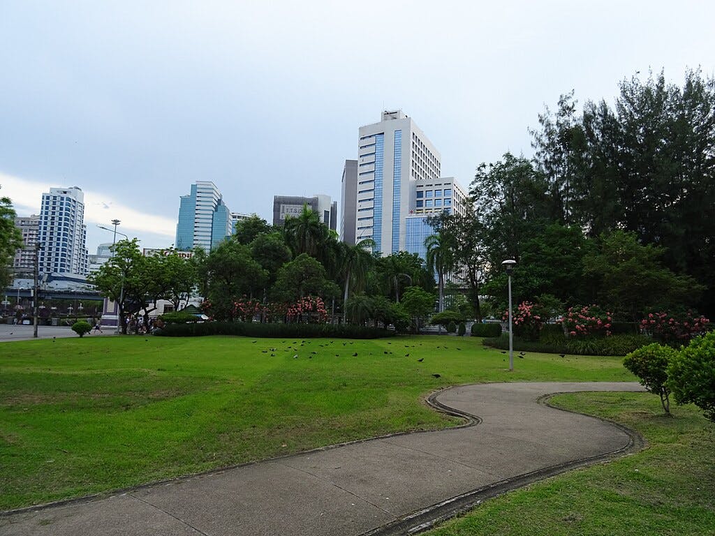 Green park with a curved concrete pathway, surrounding trees and bushes, and tall buildings in the background under a cloudy sky.