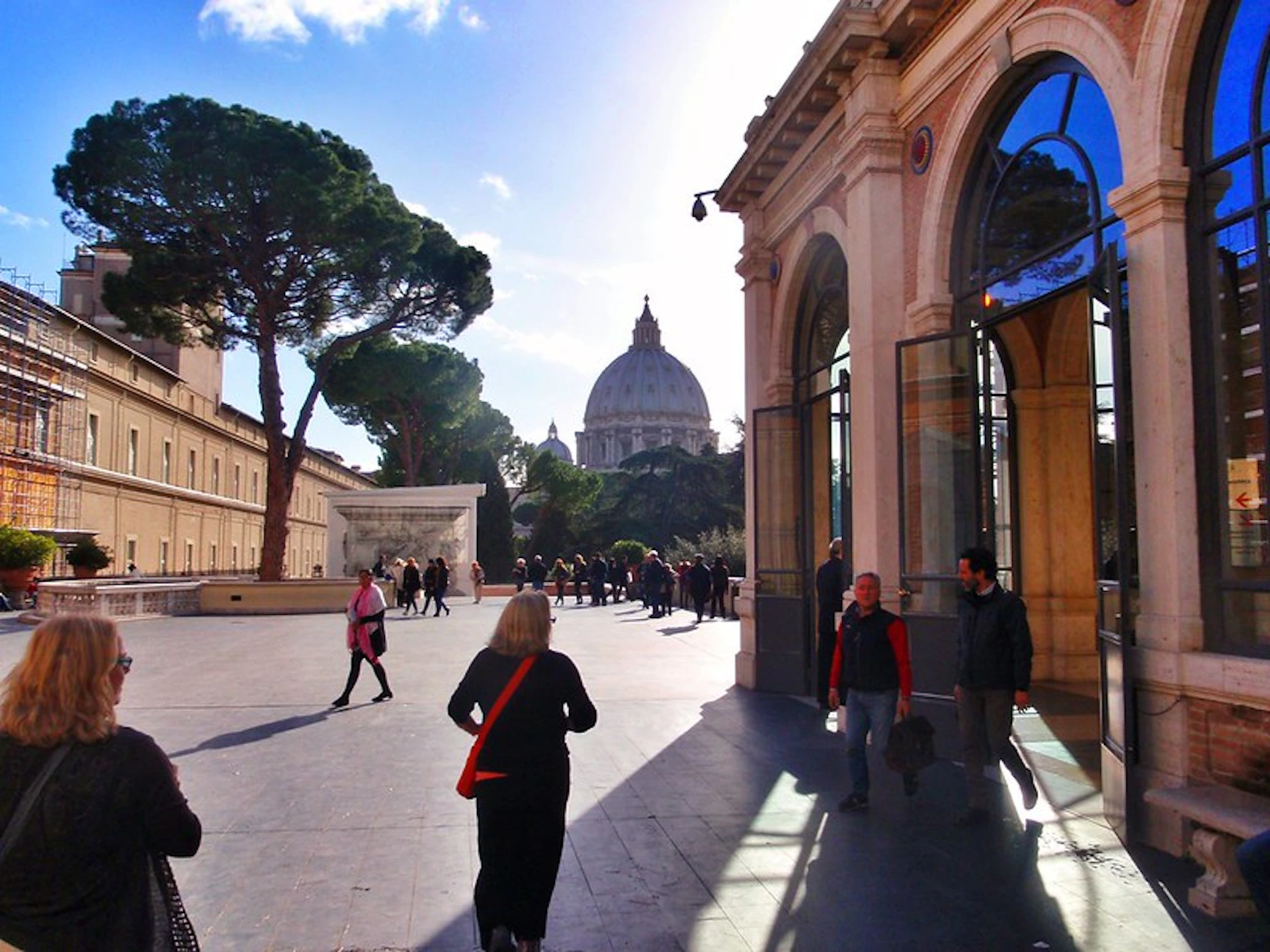 People walking in a courtyard with the dome of St. Peter's Basilica in the background under a sunny sky.
