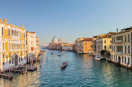 Un canale di Venezia con barche, costeggiato da edifici su entrambi i lati, sotto un cielo azzurro e limpido. La struttura a cupola è visibile sullo sfondo.