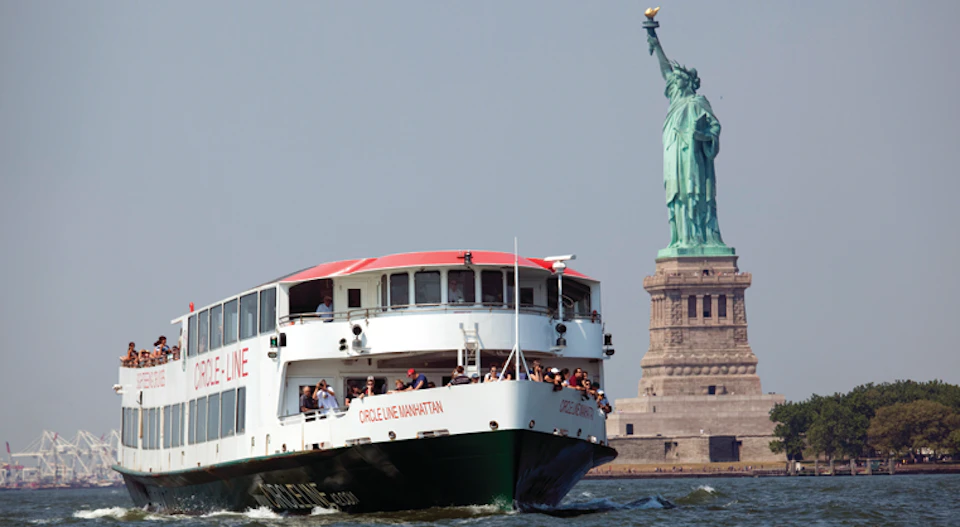 A boat with "Circle Line Manhattan" written on it passes by the Statue of Liberty on a clear day.
