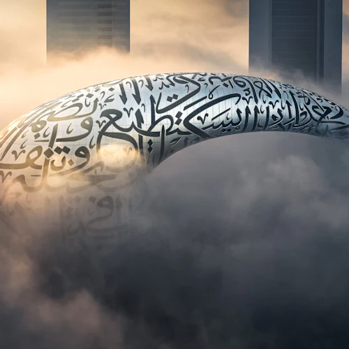 A futuristic ring-shaped building covered in Arabic calligraphy emerges through dense fog, with skyscrapers in the background.