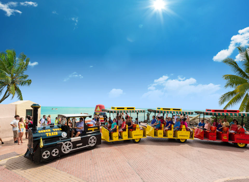 People riding a small tourist train with yellow carriages near a beach on a sunny day, with a blue sky and ocean in the background.
