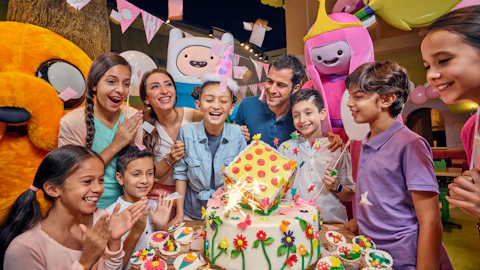 Group of children and adults celebrating a birthday with a colorful cake, cupcakes, decorations, and cartoon character mascots.