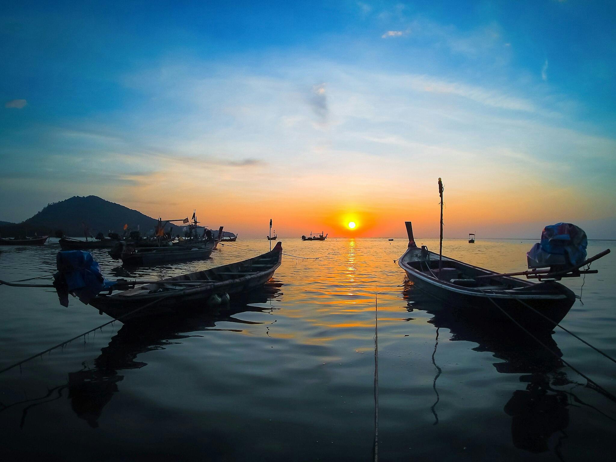 Deux bateaux flottent sur des eaux calmes face à un coucher de soleil éclatant, avec la silhouette d'une montagne lointaine sous un ciel nocturne clair.