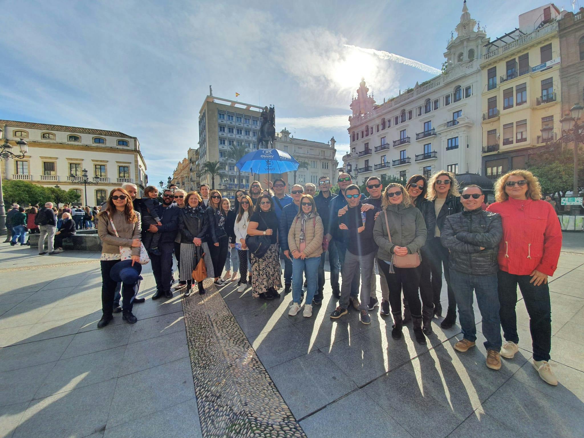 A group of people posing outdoors in front of buildings under a bright sky, with one individual holding a blue umbrella.