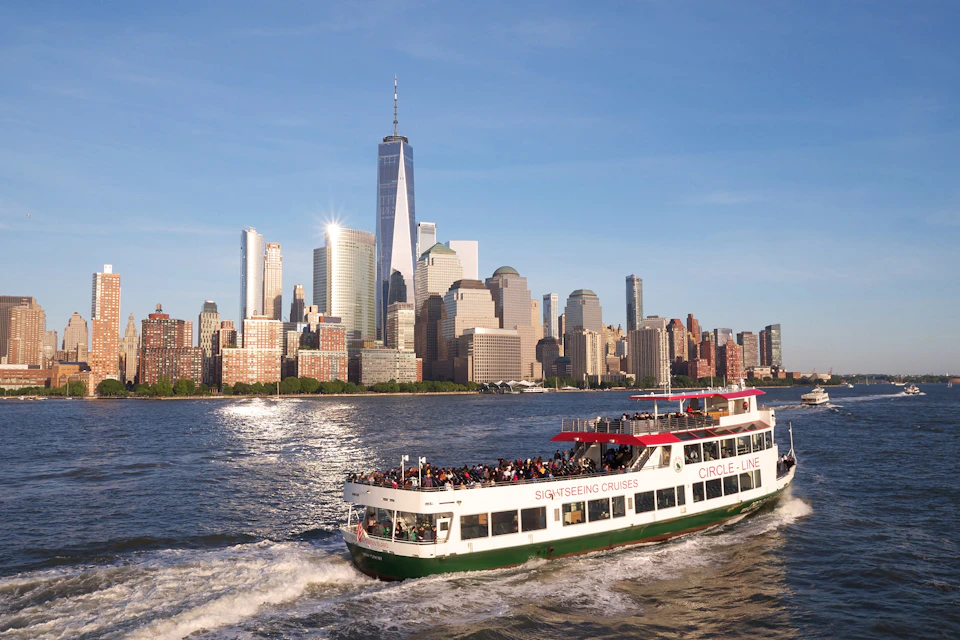 A sightseeing cruise boat on the water with a group of people on board, with the New York City skyline in the background.
