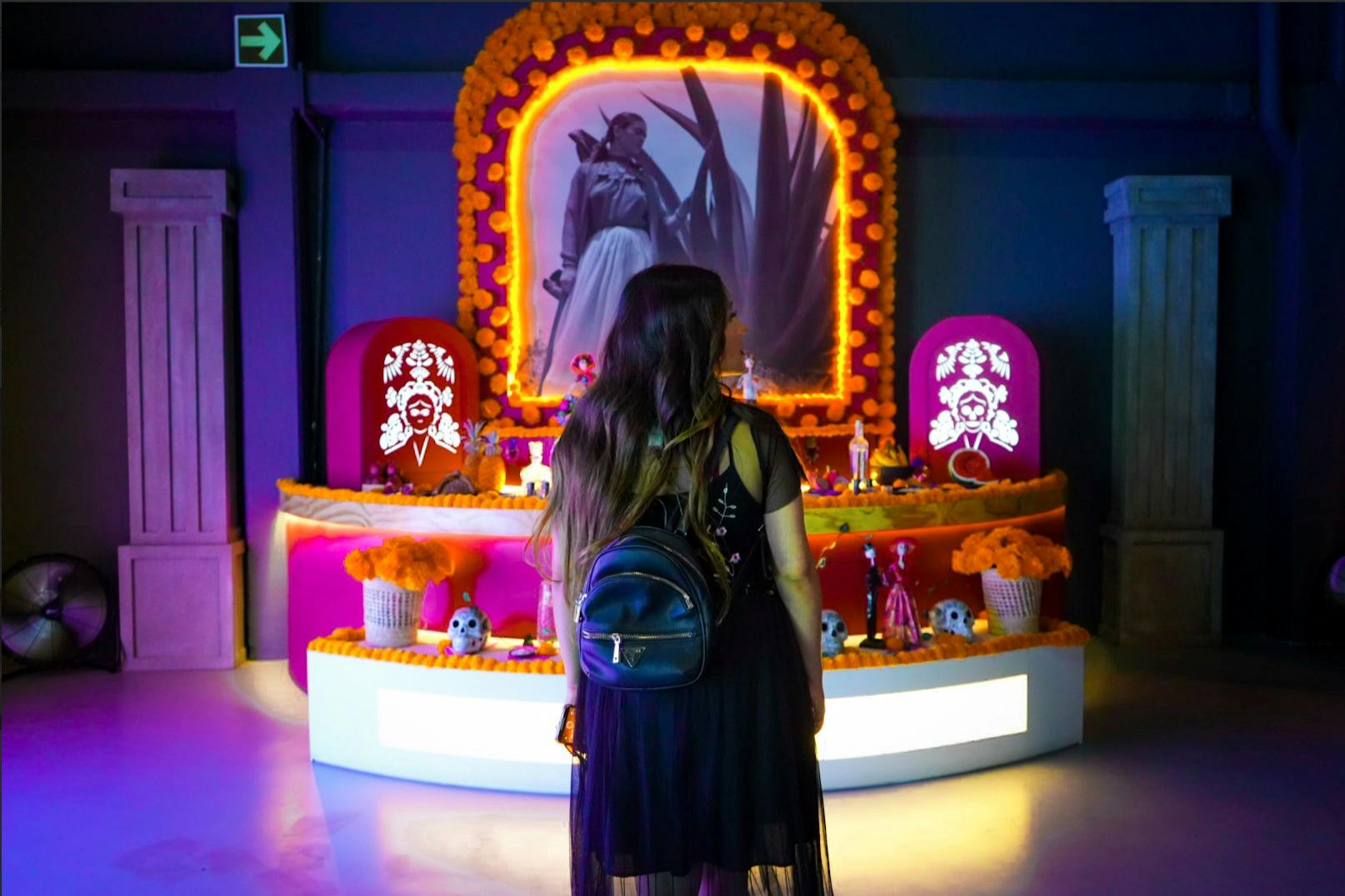 Μια γυναίκα με σακίδιο στέκεται μπροστά από έναν διακοσμημένο βωμό της Día de los Muertos με κατιφέδες, κεριά και φωτογραφίες σε ένα αμυδρά φωτισμένο δωμάτιο.