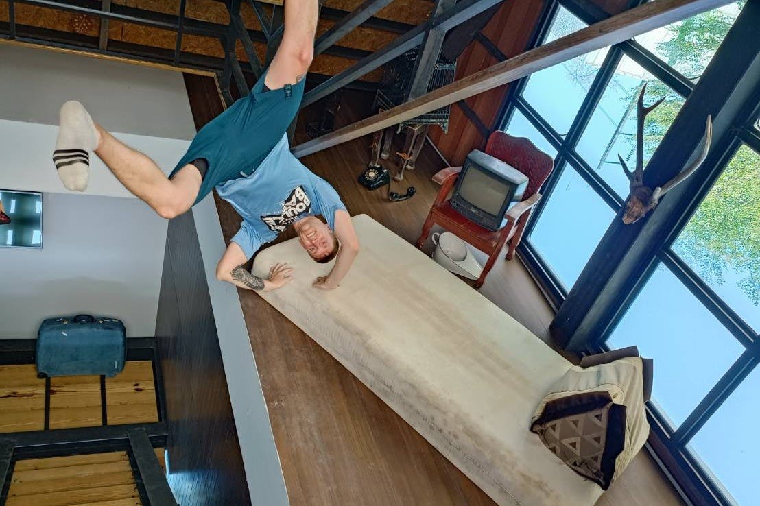 Un hombre cuelga cabeza abajo de la barandilla de un altillo sobre una habitación con una cama, un televisor antiguo, una silla y ventanas con vistas a los árboles.