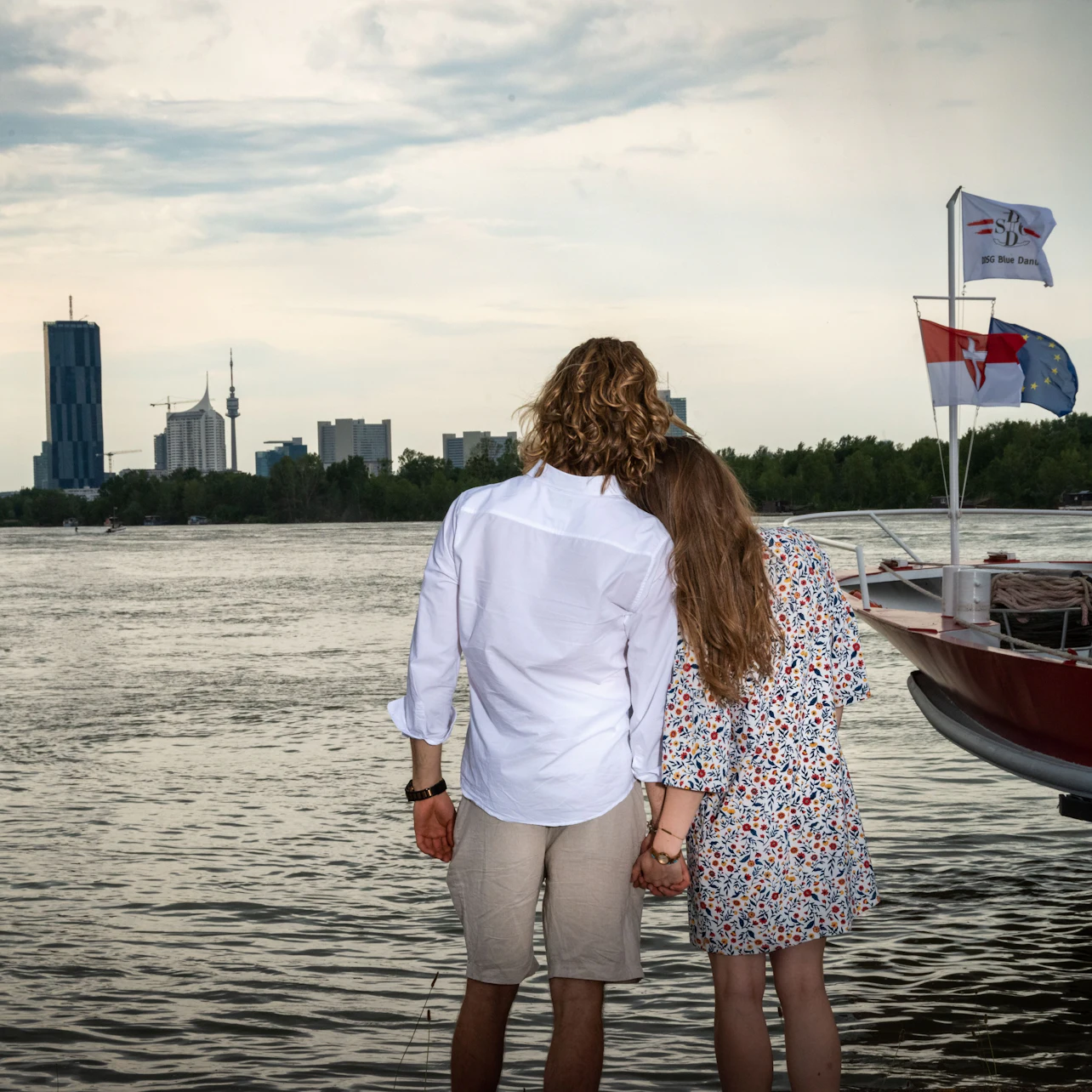 A couple holds hands, facing a river with a boat and city skyline in the background, featuring flags and tall buildings.