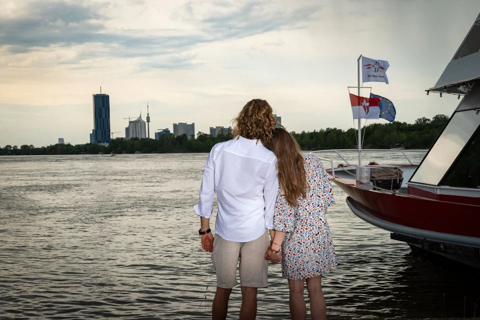 A couple holds hands, facing a river with a boat and city skyline in the background, featuring flags and tall buildings.