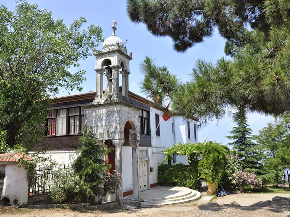 A small white building with an arched entrance, bell tower, and stairs, surrounded by trees and greenery under a clear sky.