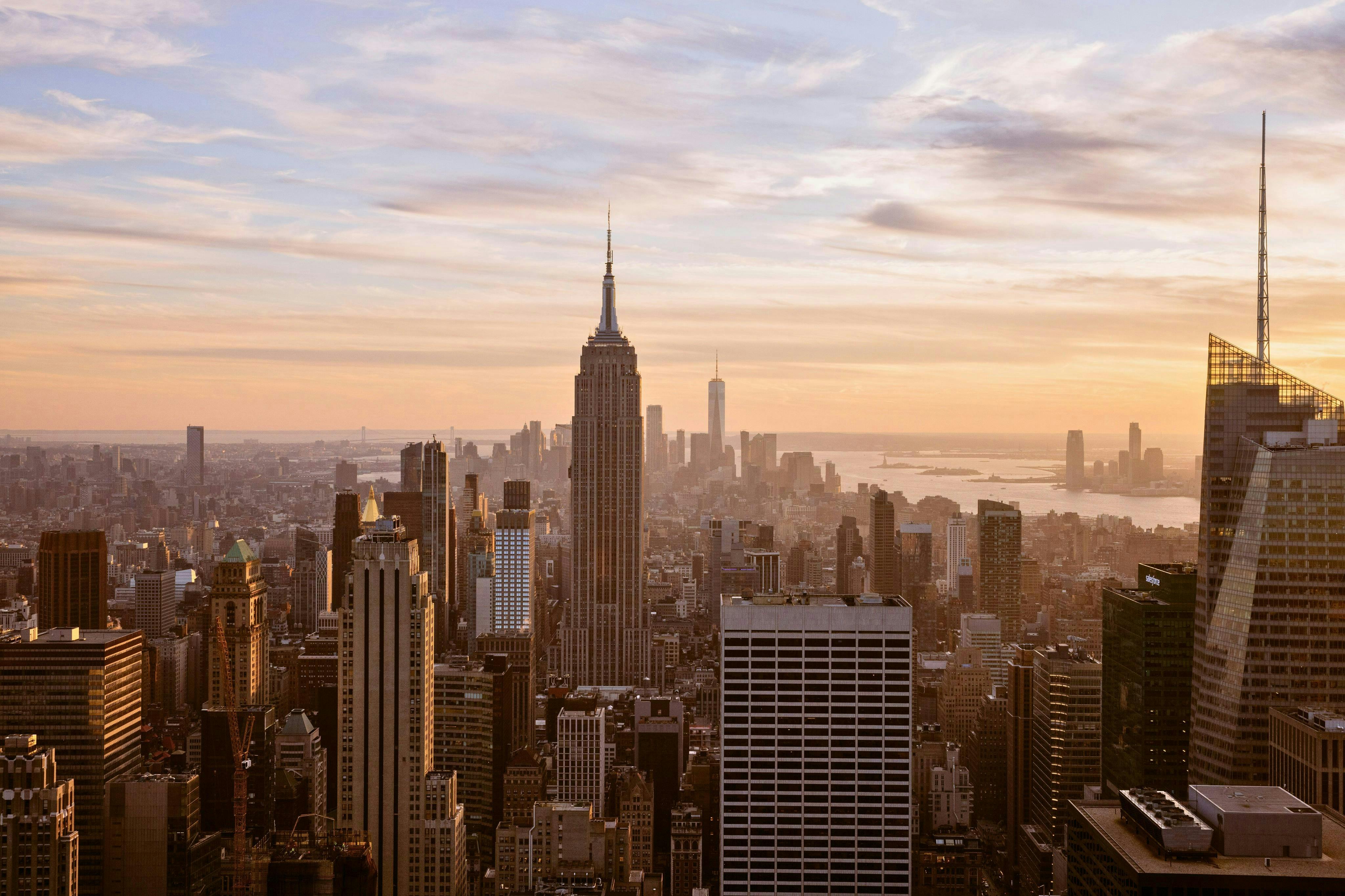 Uma paisagem urbana com o Empire State Building ao pôr-do-sol, rodeado por arranha-céus e um céu dourado.