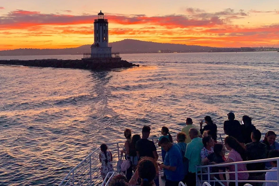 A group of people on a boat watch a sunset over a lighthouse on a rocky outcrop with hills in the background.