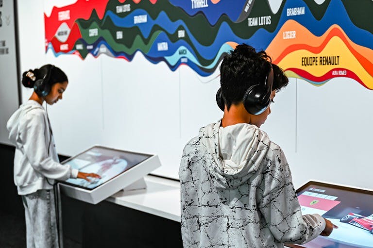 Els nens interactuen amb una pantalla tàctil en una exposició amb auriculars. A la paret es pot veure un colorit gràfic ondulat.