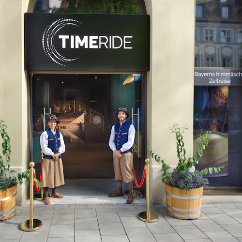 Zwei Personen in traditioneller Kleidung stehen vor dem Eingang von Timeride, einem Virtual-Reality-Erlebniszentrum.