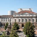 Historic building with ornate facade and statue in foreground, surrounded by greenery and people sitting on benches.