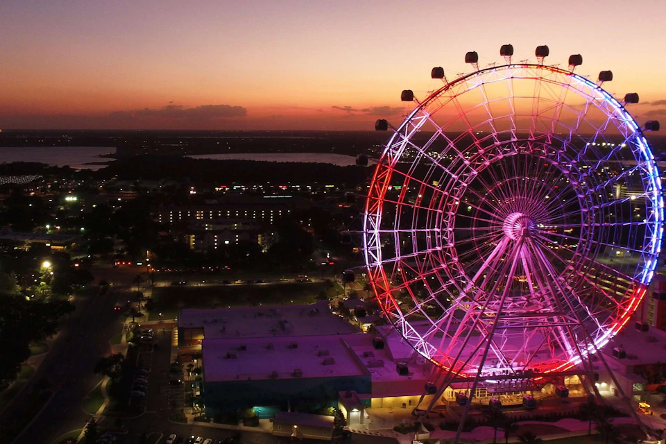 A large illuminated Ferris wheel at dusk, with a cityscape and a lake in the background.