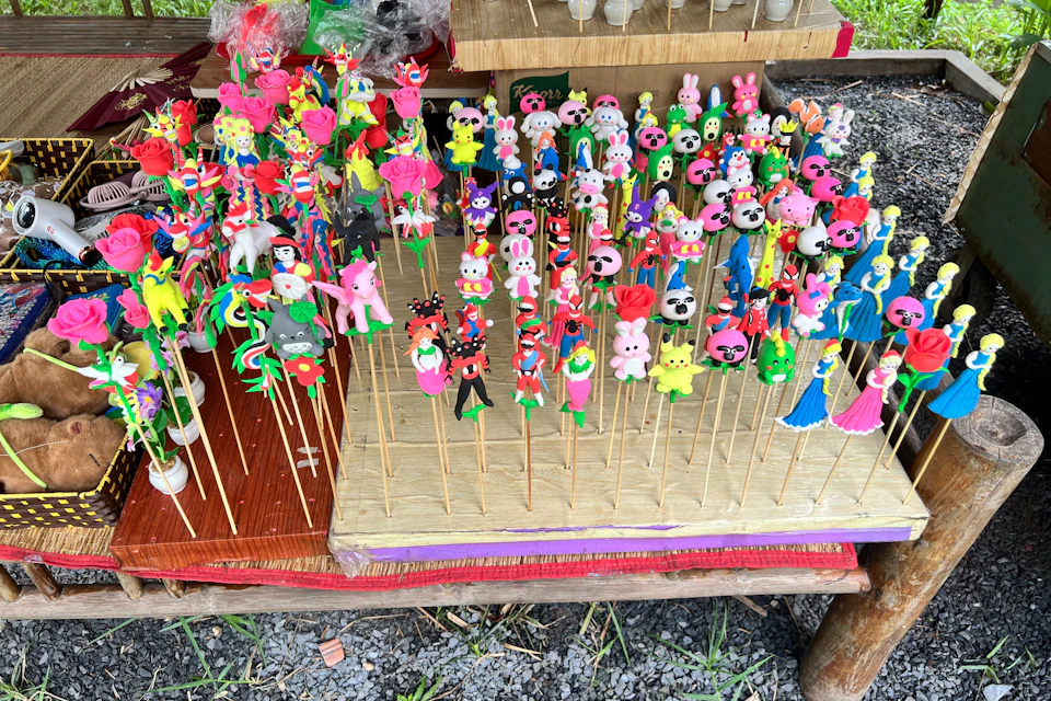 Colorful clay figurines on sticks displayed on wooden boards at an outdoor market, featuring various characters and animals.