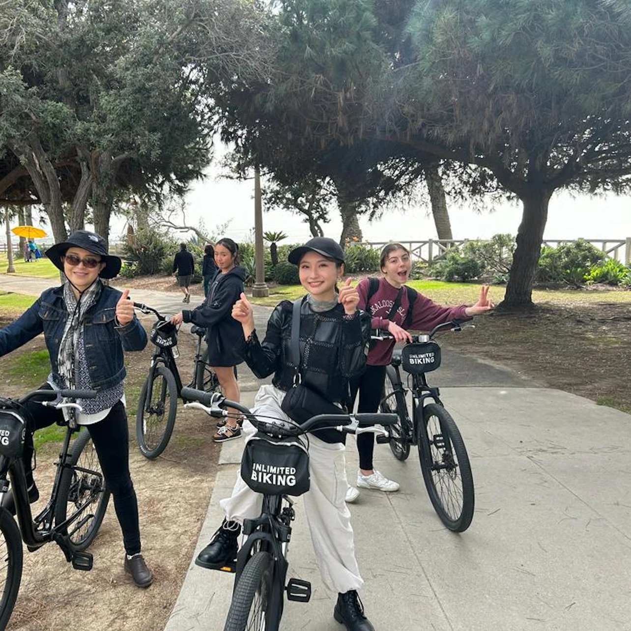 Four people pose with rental bikes on a tree-lined pathway near a park. They are smiling and giving thumbs-up gestures.