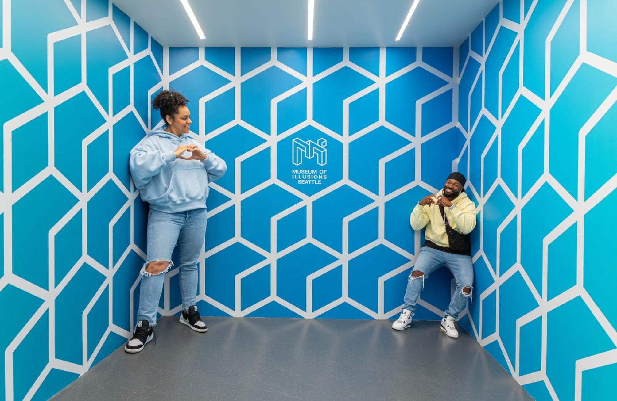 Eine Person in Freizeitkleidung steht in einem Raum mit blauen geometrischen Wandmustern und der Aufschrift "Museum of Illusions Seattle".