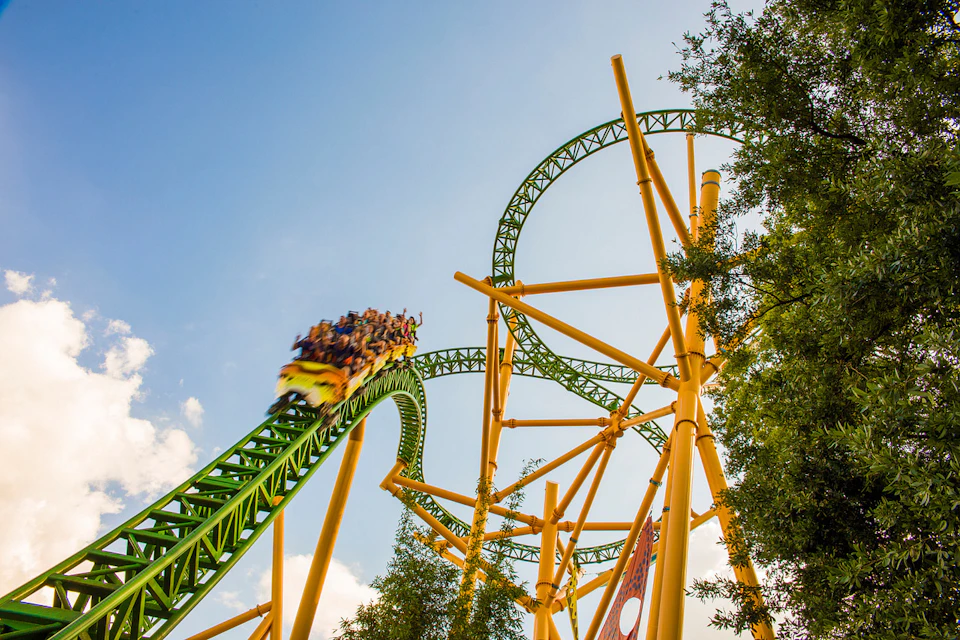 Rollercoaster with green tracks and yellow supports, full of people, ascending a steep curve under a clear blue sky.