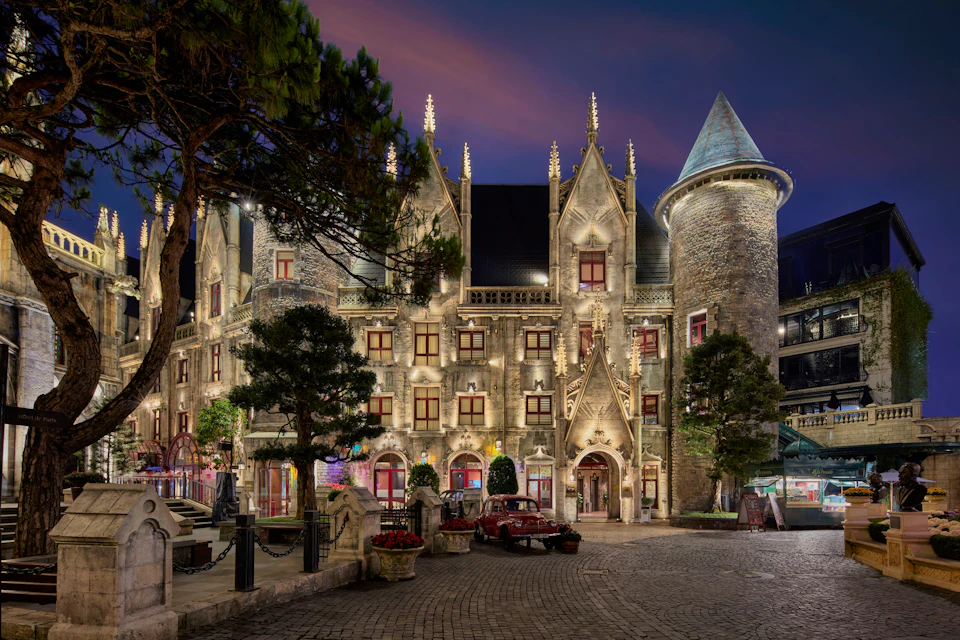 An illuminated castle-like building with towers and spires at night, featuring a cobblestone courtyard and vintage car.