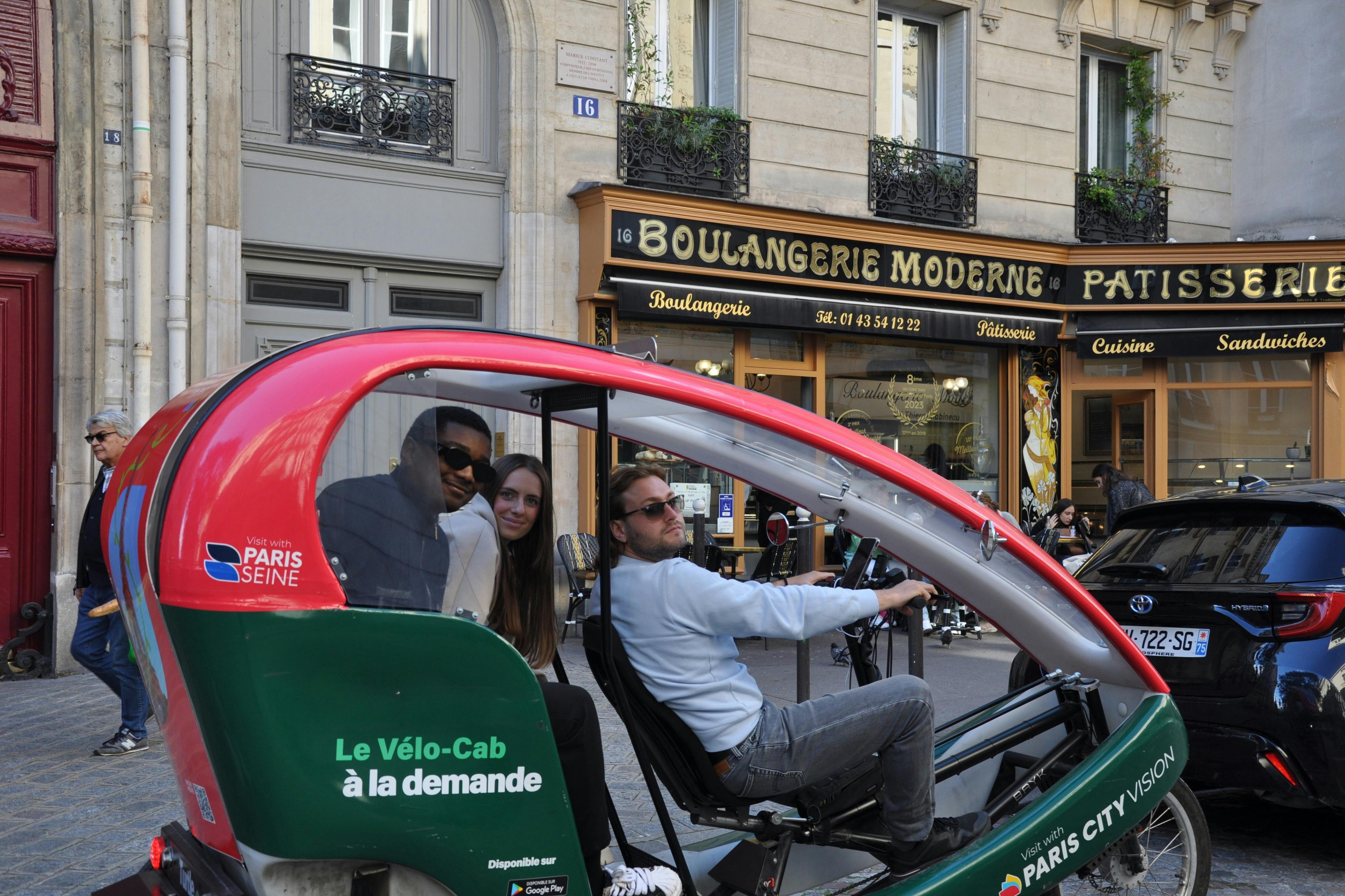 Three people riding in a green and red pedicab in front of a bakery with "Boulangerie Moderne" signage.