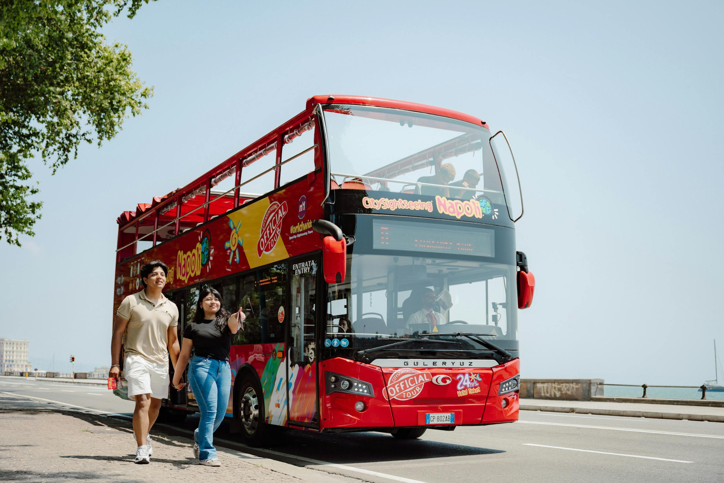 Una coppia cammina vicino a un autobus turistico rosso a due piani parcheggiato su una strada sotto un cielo limpido con alcuni alberi in vista.