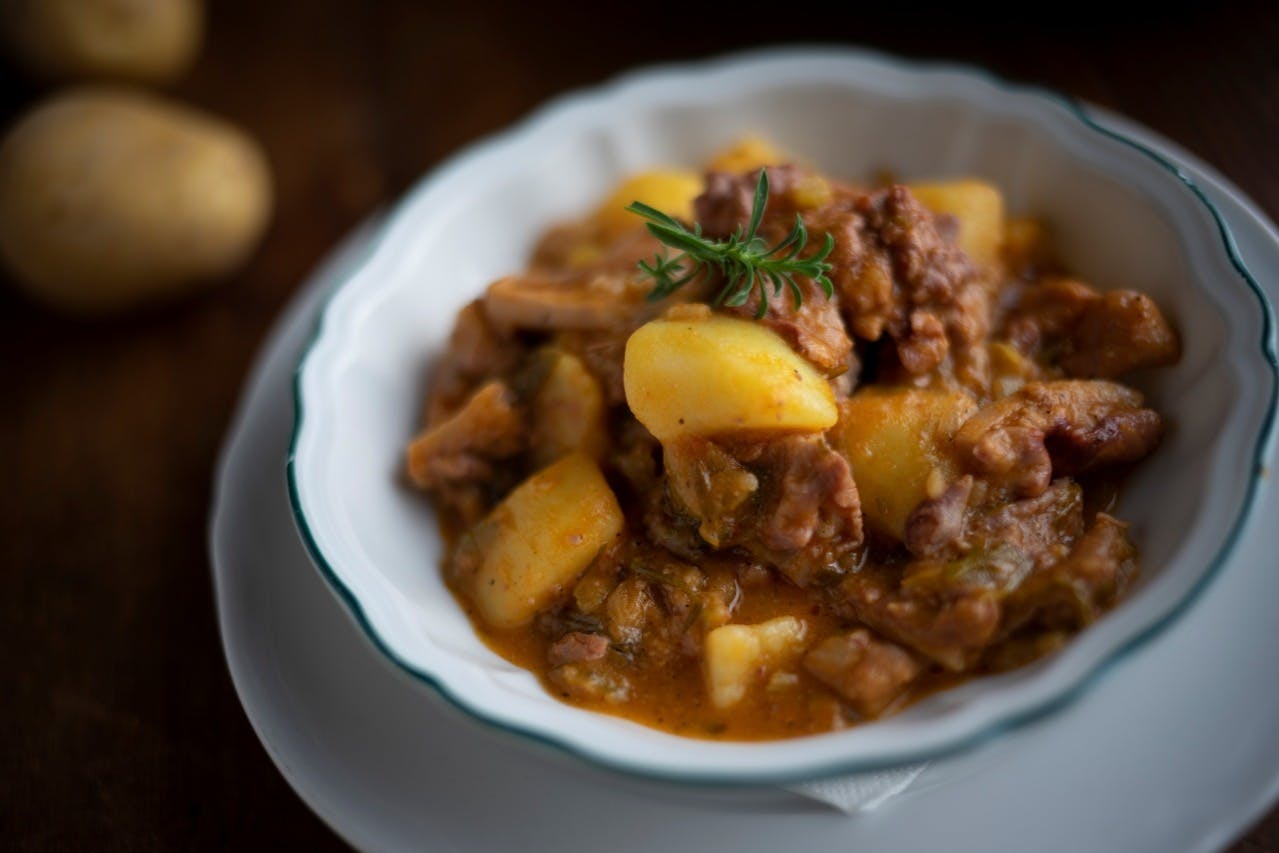 A white bowl filled with a hearty stew of potatoes, meat, and garnished with a sprig of rosemary.