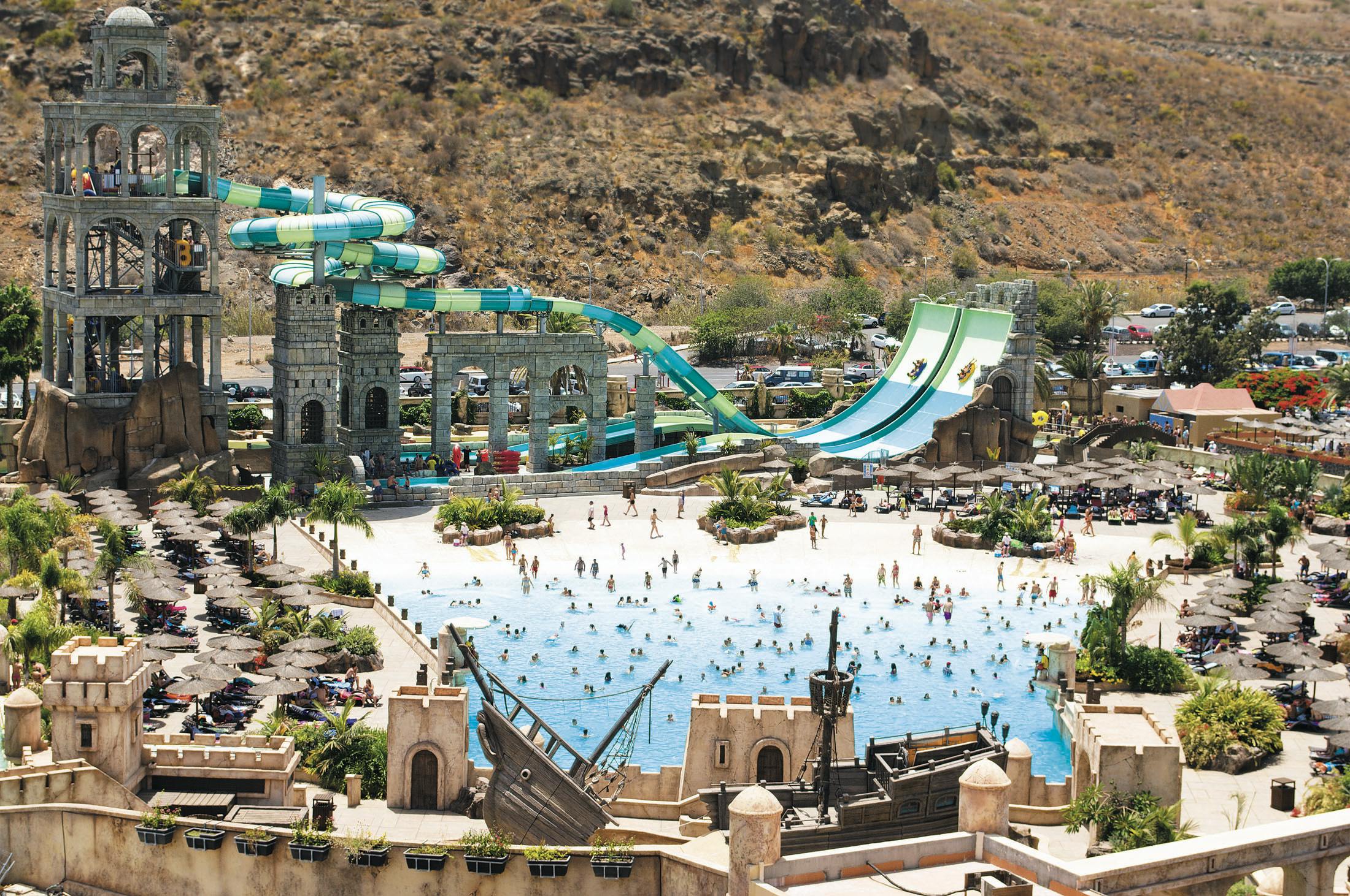 A water park with a wave pool, large slides, and a pirate ship structure surrounded by people enjoying the attractions.