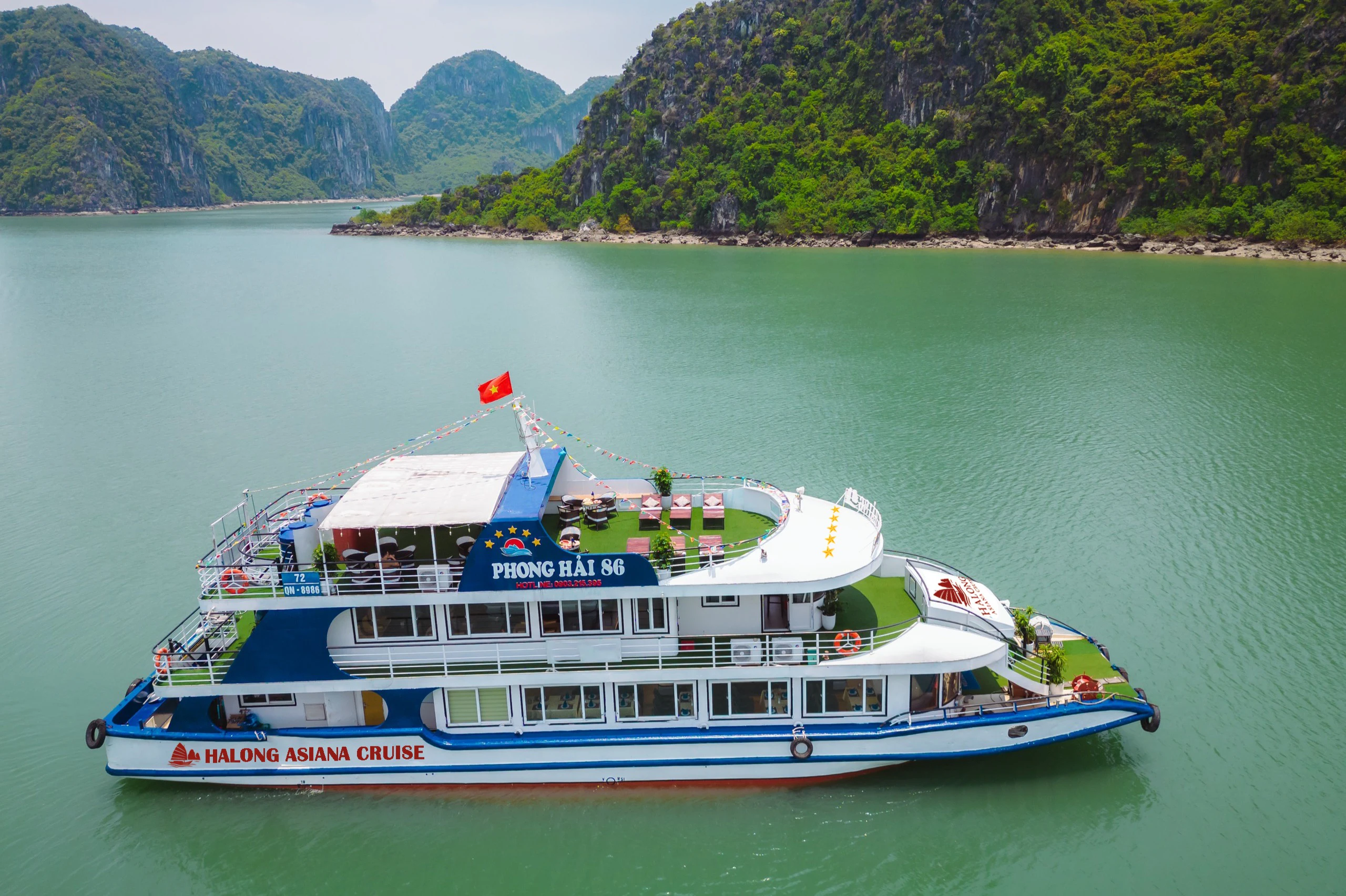 White and blue cruise boat on calm green water, near lush, rocky cliffs under a clear sky. Various seating areas on the decks.