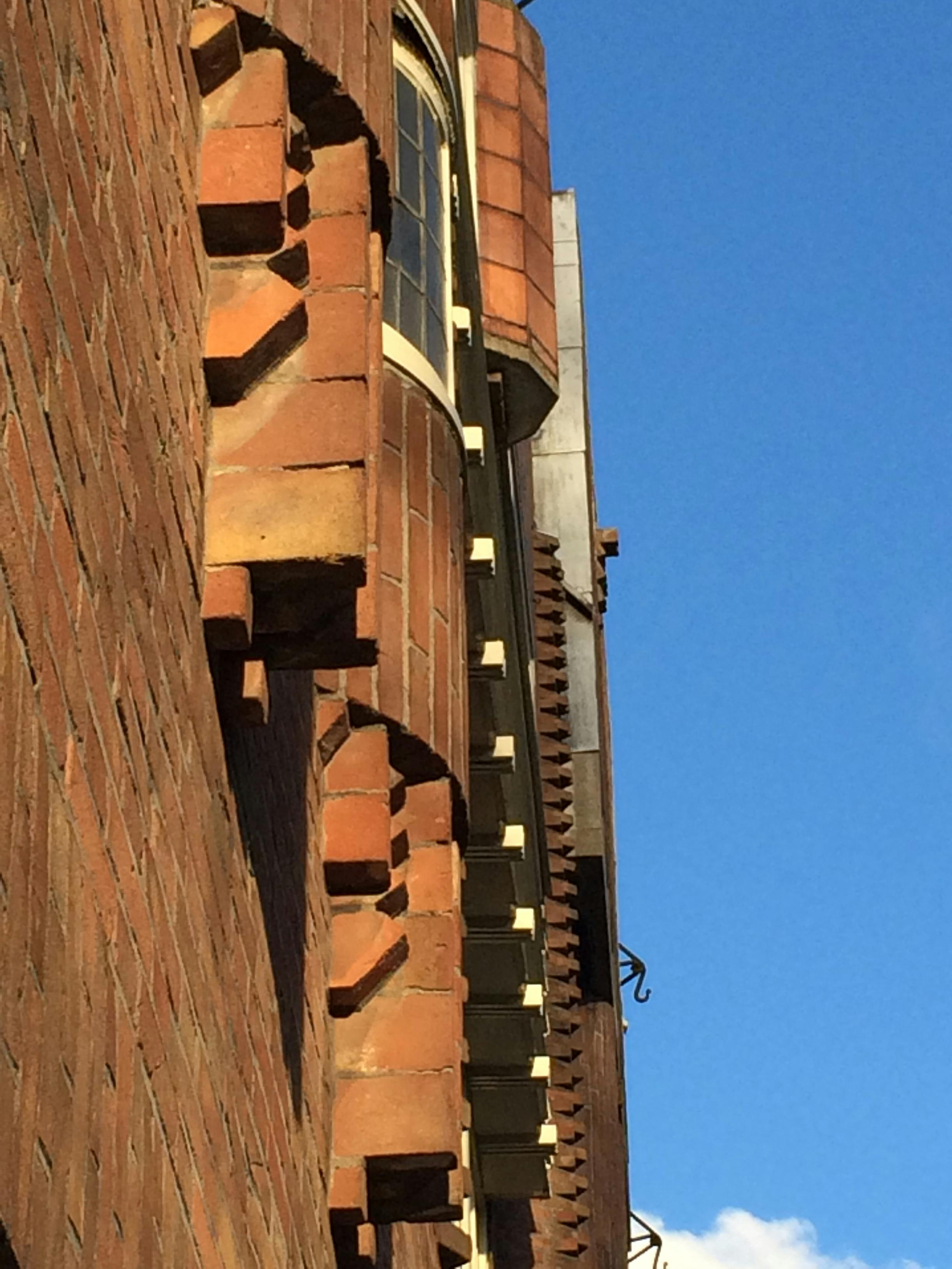 Budynek z czerwonej cegły z wystającymi detalami murowanymi, częściowo widocznym oknem i czystym błękitnym niebem.