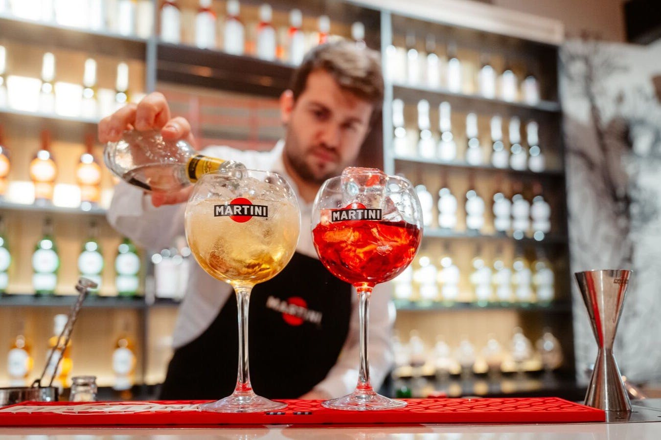 A bartender pours drinks into two glasses labeled "Martini" at a bar with shelves of bottles in the background.