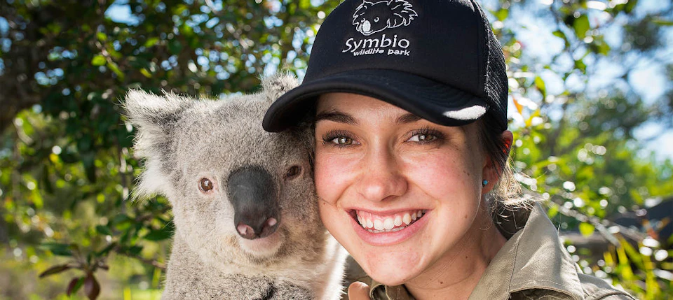 A smiling woman wearing a "Symbio Wildlife Park" cap stands close to a koala, with greenery in the background.
