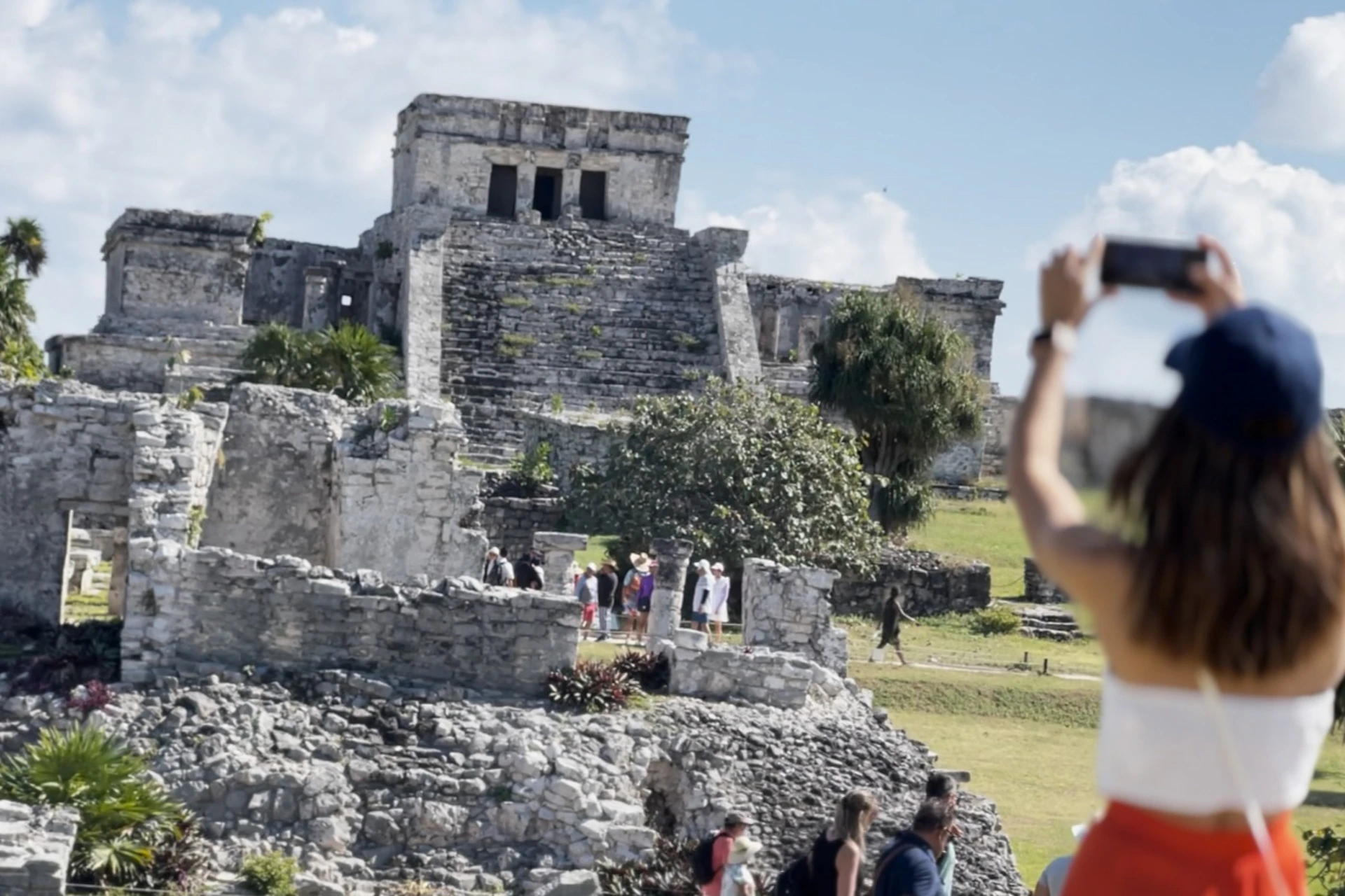 Tourists explore an ancient stone ruin under a clear sky, with one person taking a photo in the foreground.
