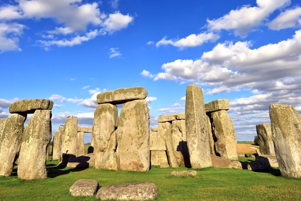 Stonehenge under a blue sky with scattered clouds, surrounded by green grass.