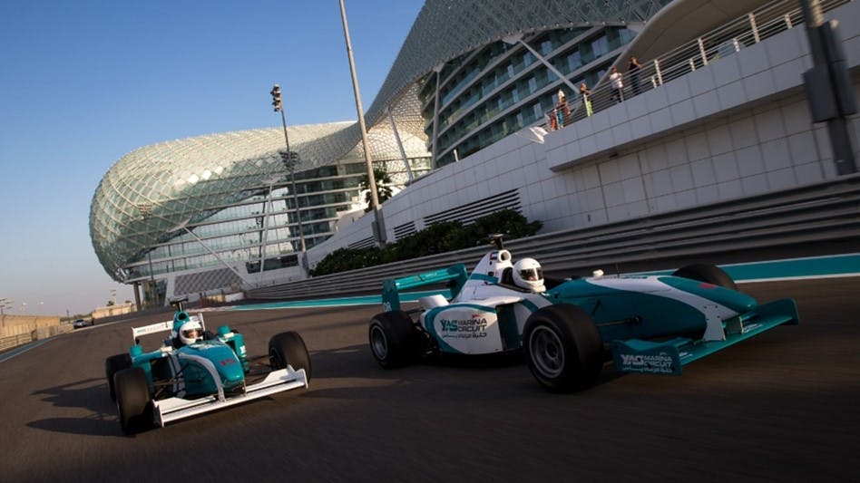 Two race cars on a track with a modern, lattice-covered building and spectators in the background under a clear blue sky.