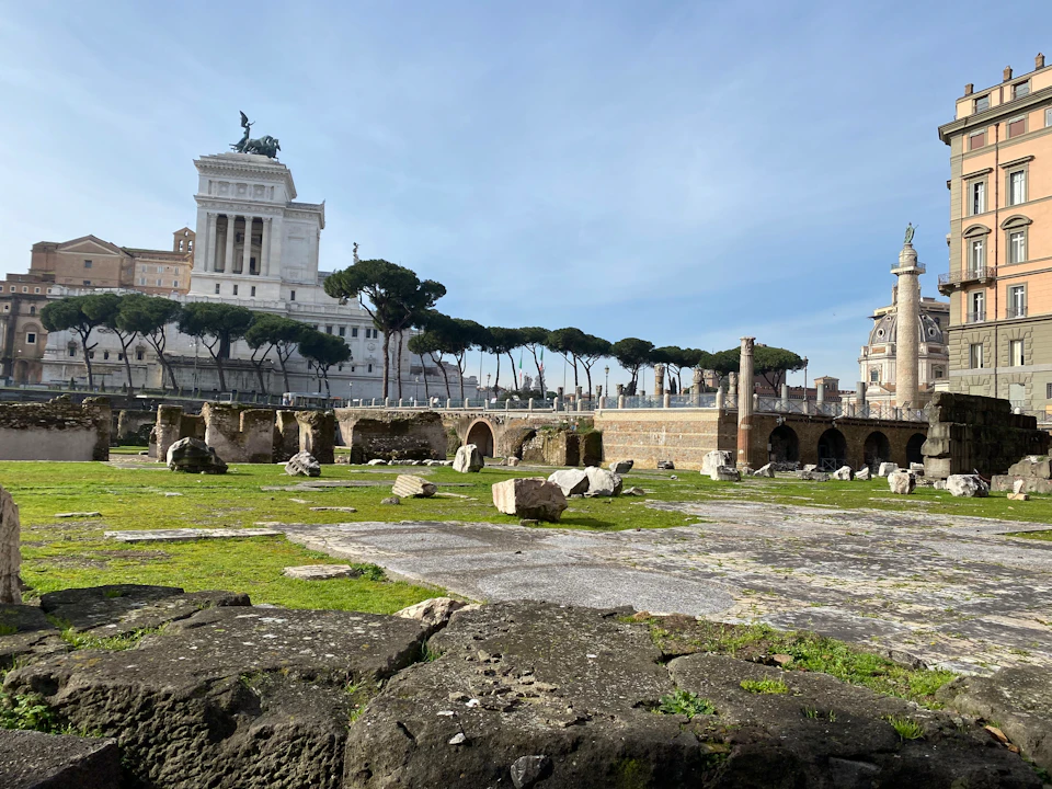 View of ancient ruins with scattered stone remnants, a backdrop of arches, and a large monument with a statue on top in the distance.