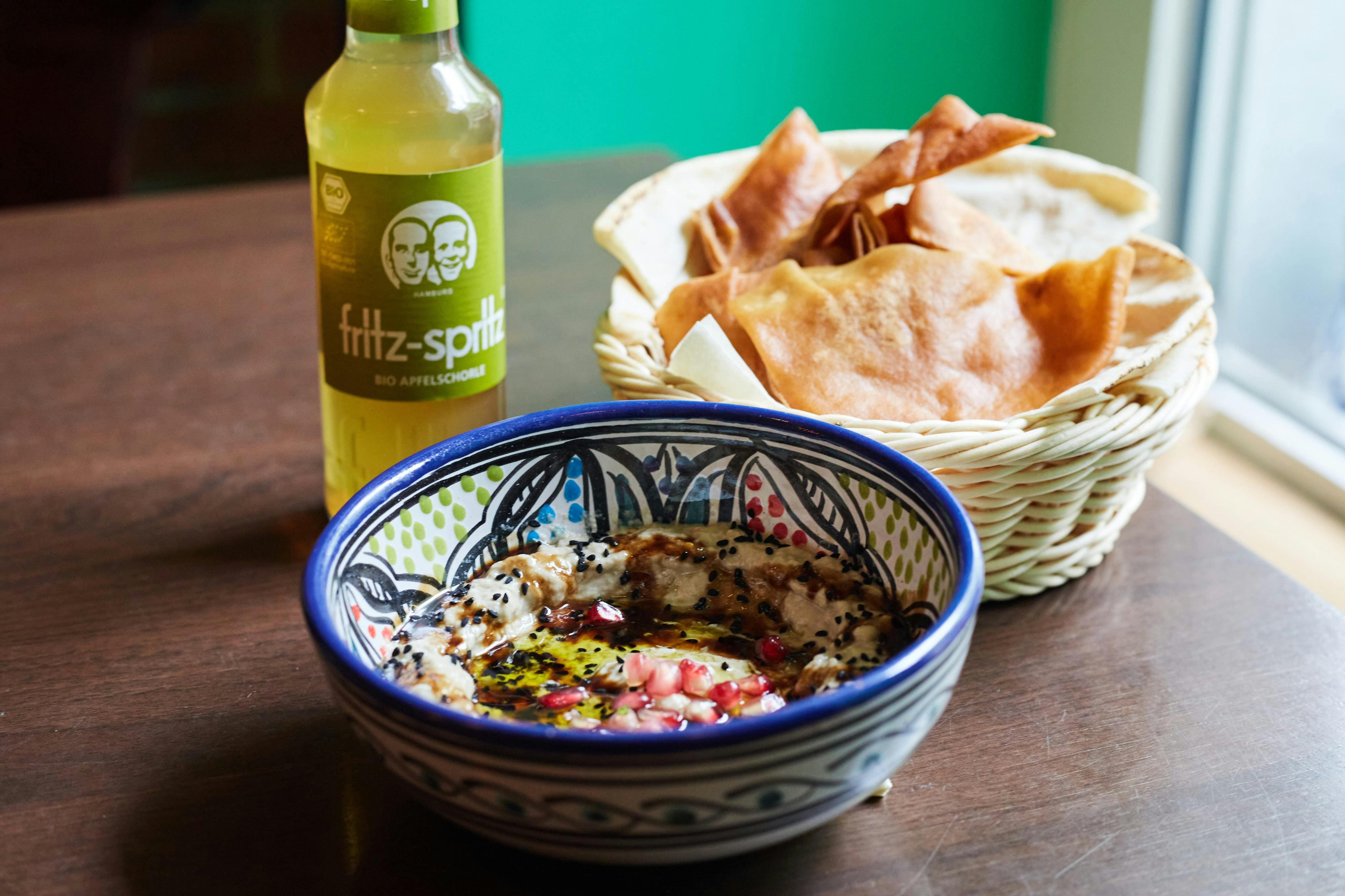 Ένα μπολ χούμους γαρνιρισμένο με ελαιόλαδο και σπόρους ροδιού, σερβιρισμένο με πίτα και ένα μπουκάλι Fritz-Spritz.