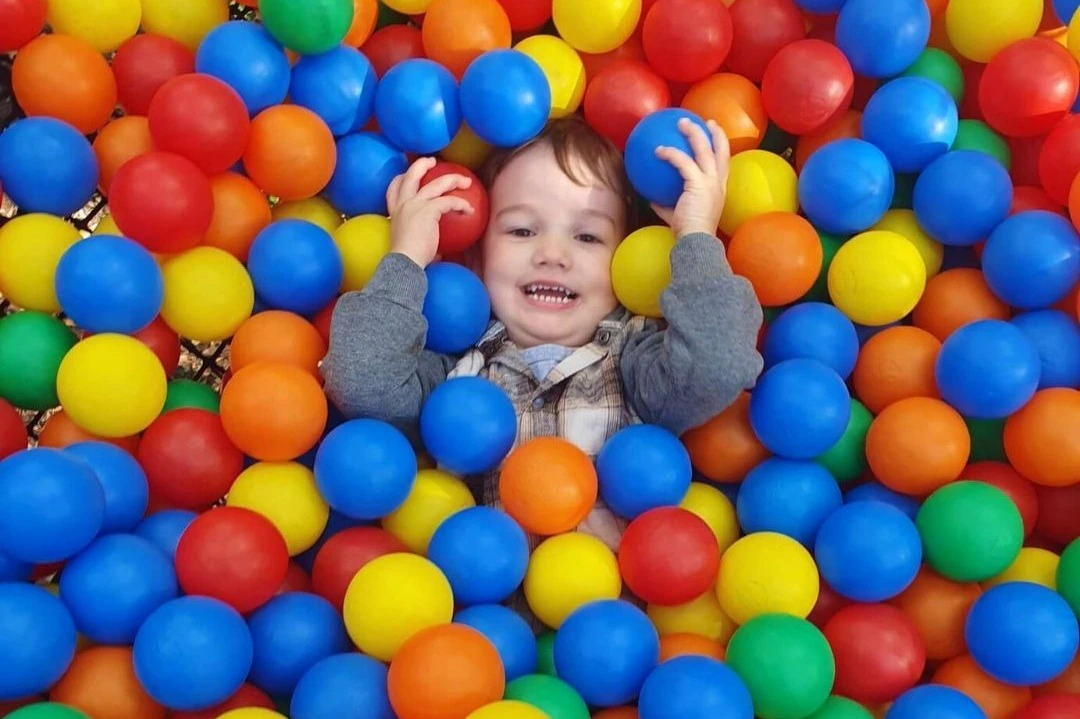 A child smiling and lying in a colorful ball pit, surrounded by red, blue, yellow, and green plastic balls.
