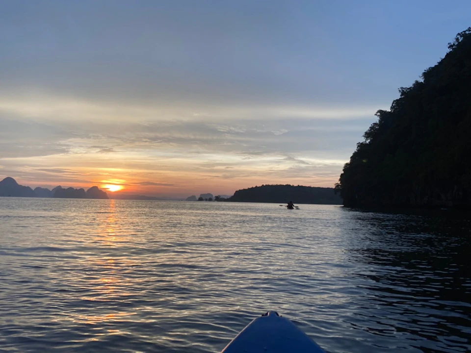 Kayak on calm water at sunset with a distant silhouette of a kayaker and shoreline against a colorful sky.