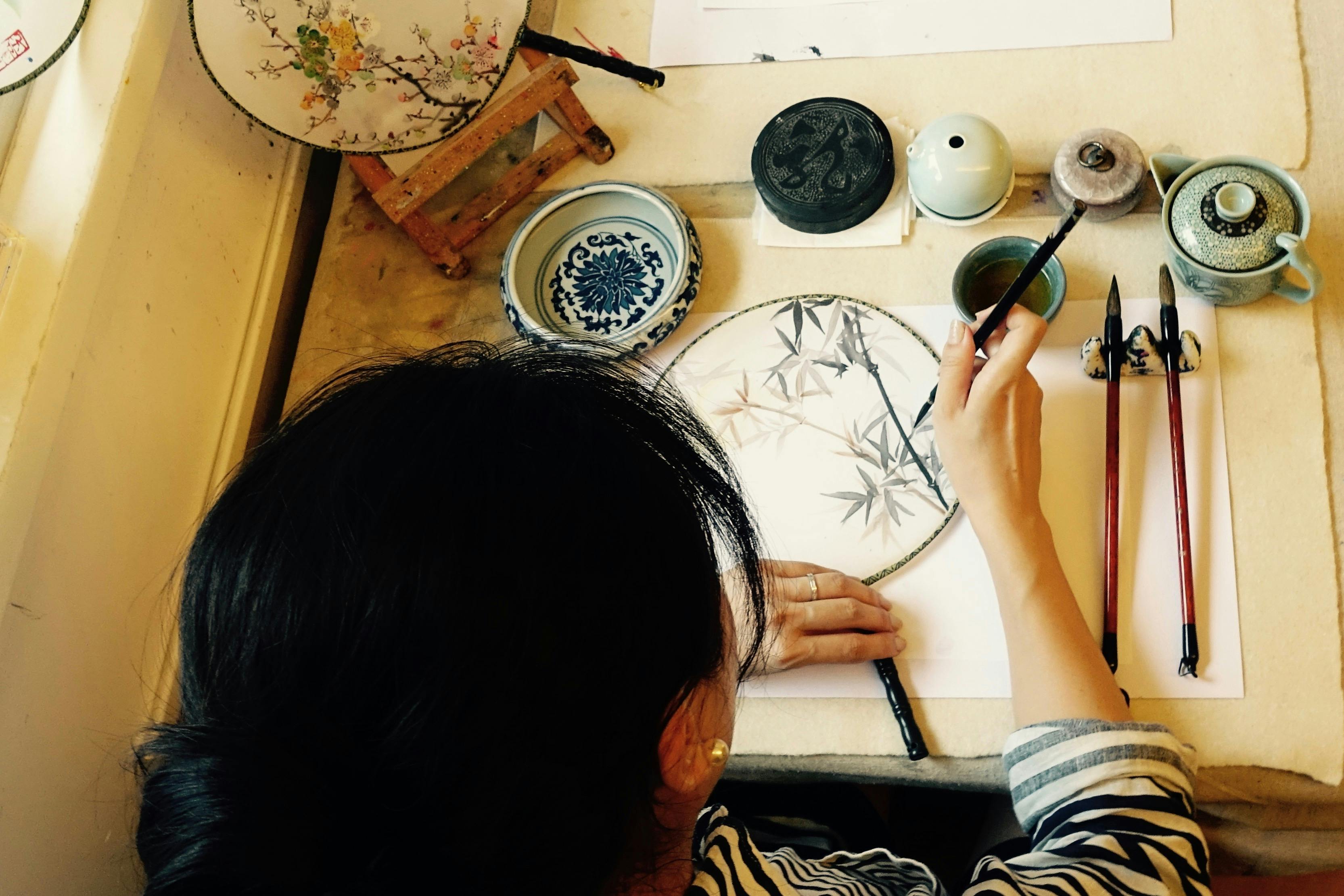 Person painting bamboo on a round fan, surrounded by traditional Chinese art supplies and ceramics.