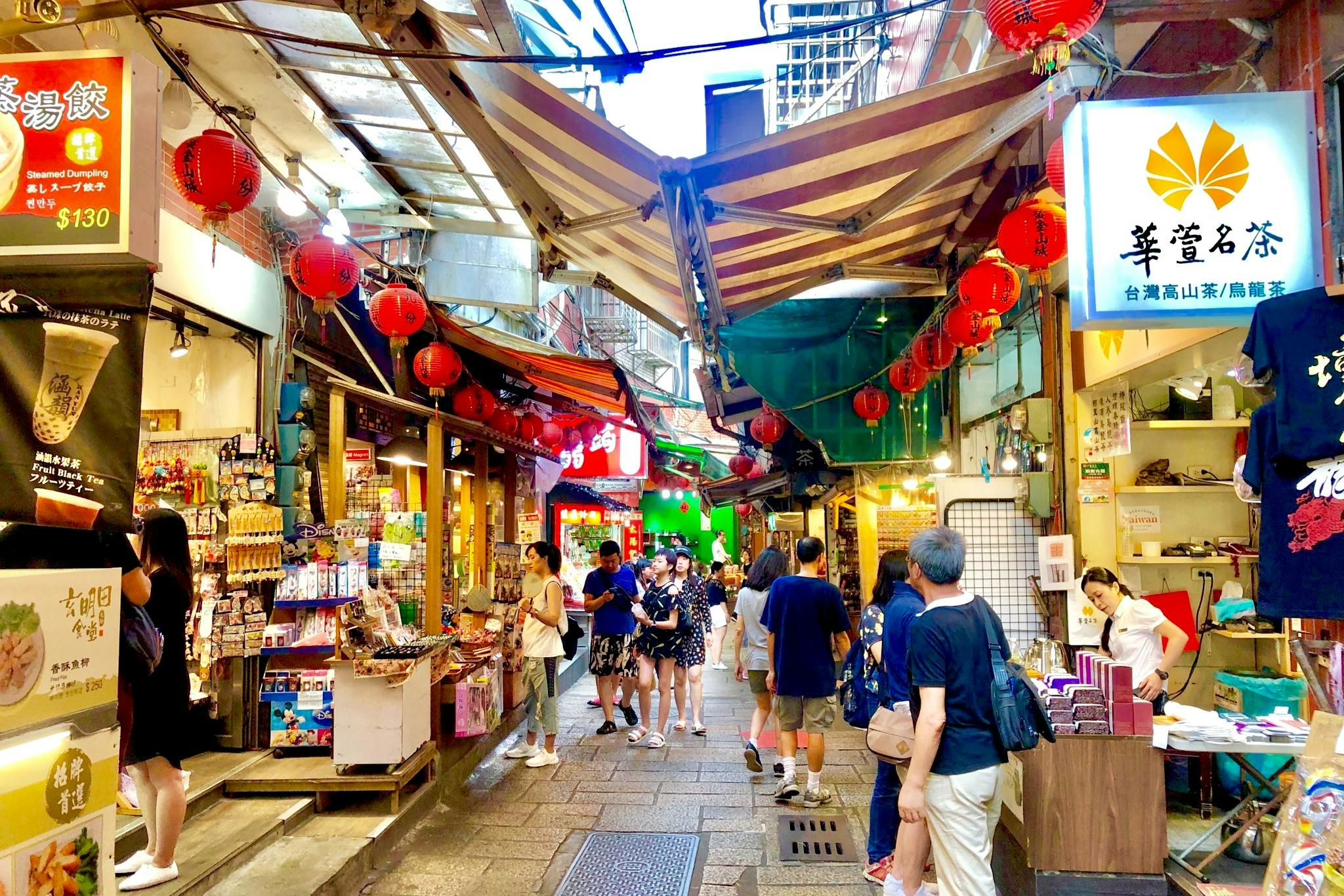 Busy market street with red lanterns, various shops, and people walking or shopping. Awnings cover the narrow pathway.