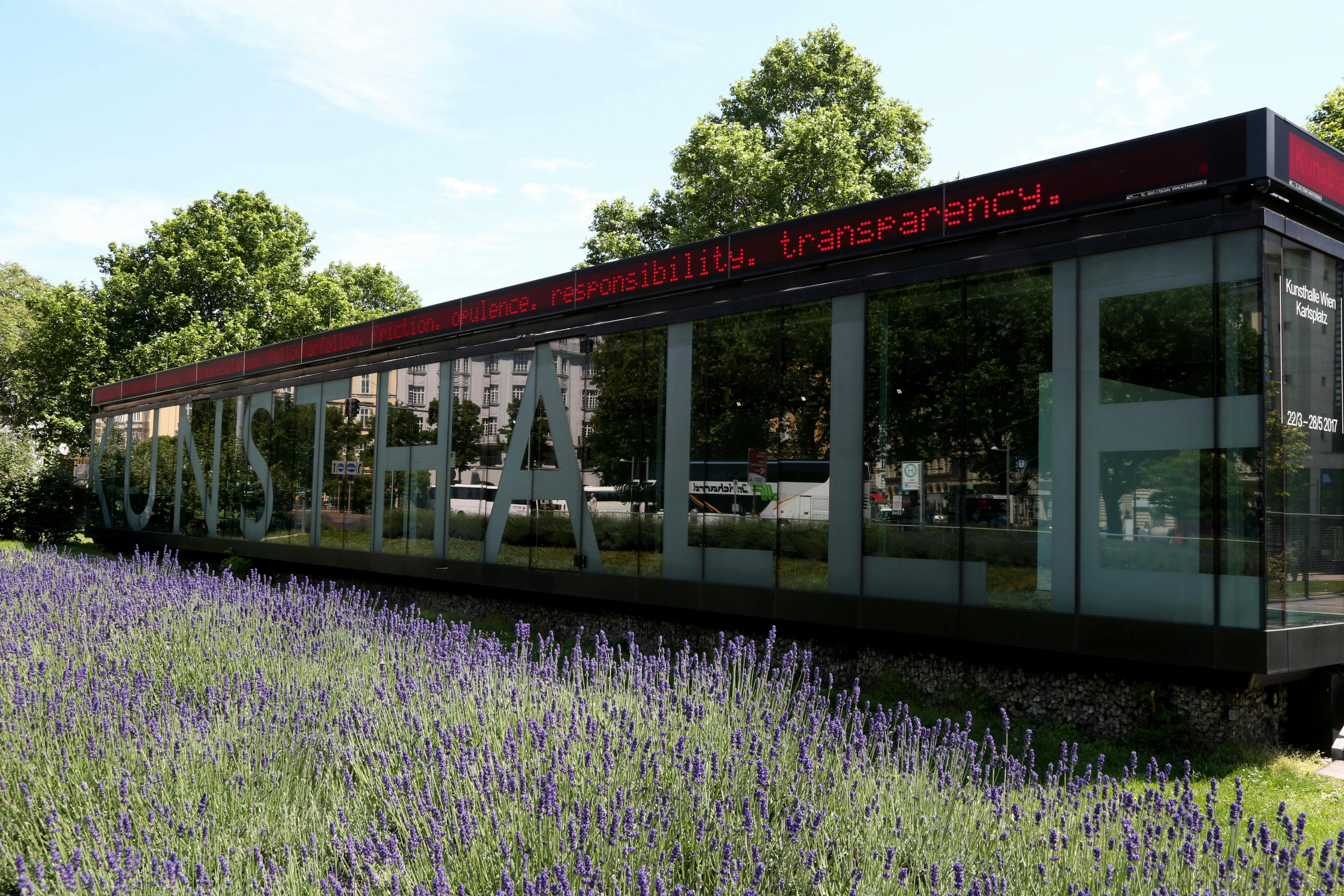 En bygning med store reflekterende vinduer, et LED-skilt over og en forgrund af blomstrende lavendelplanter.