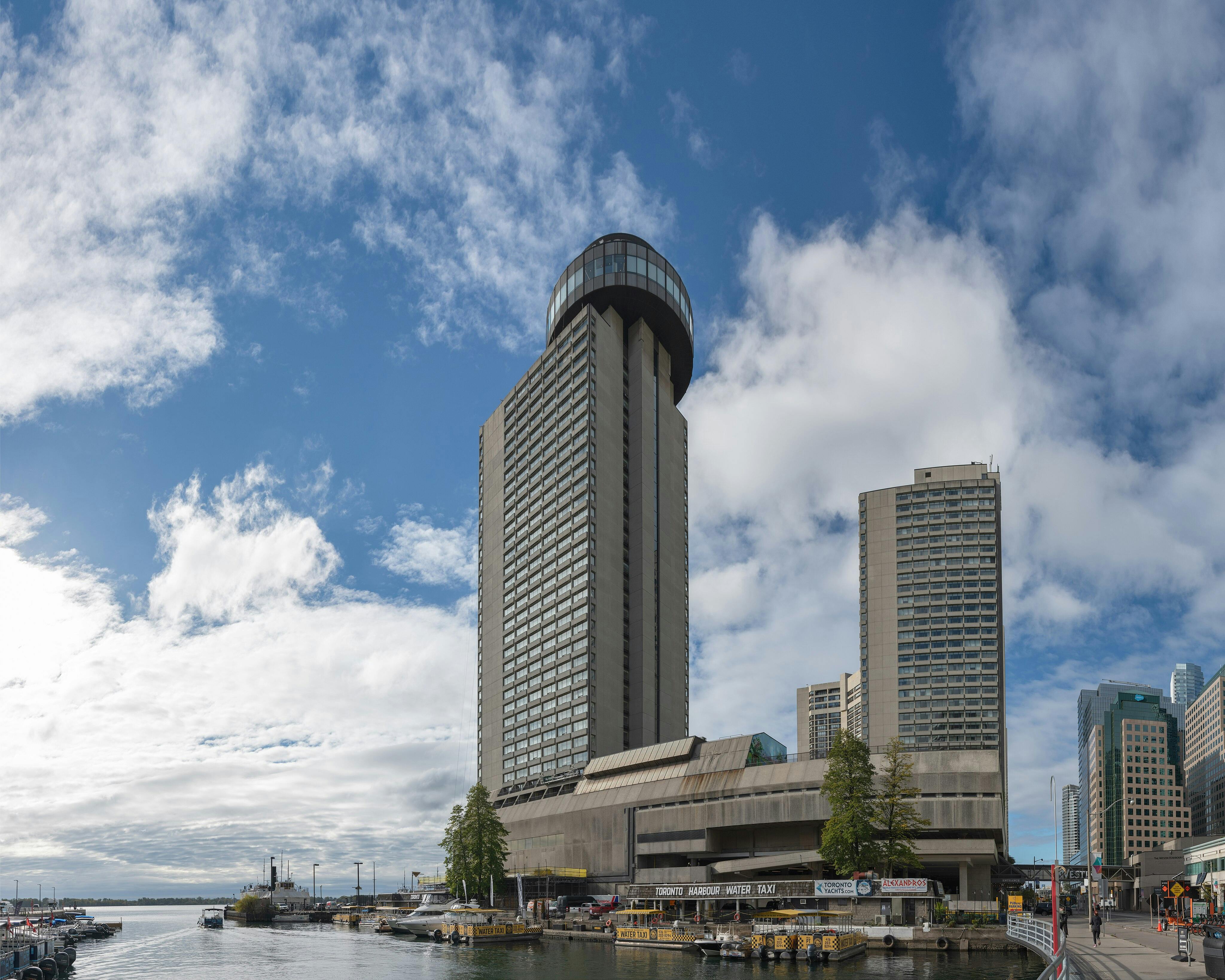 Grands immeubles près d'un front de mer par une journée partiellement nuageuse, avec des quais et des bateaux en contrebas et des personnes marchant sur un sentier à proximité.