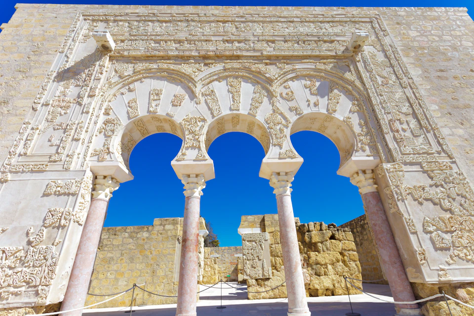 Three ornate stone arches with intricate carvings, supported by columns, against a bright blue sky and ancient stone walls.