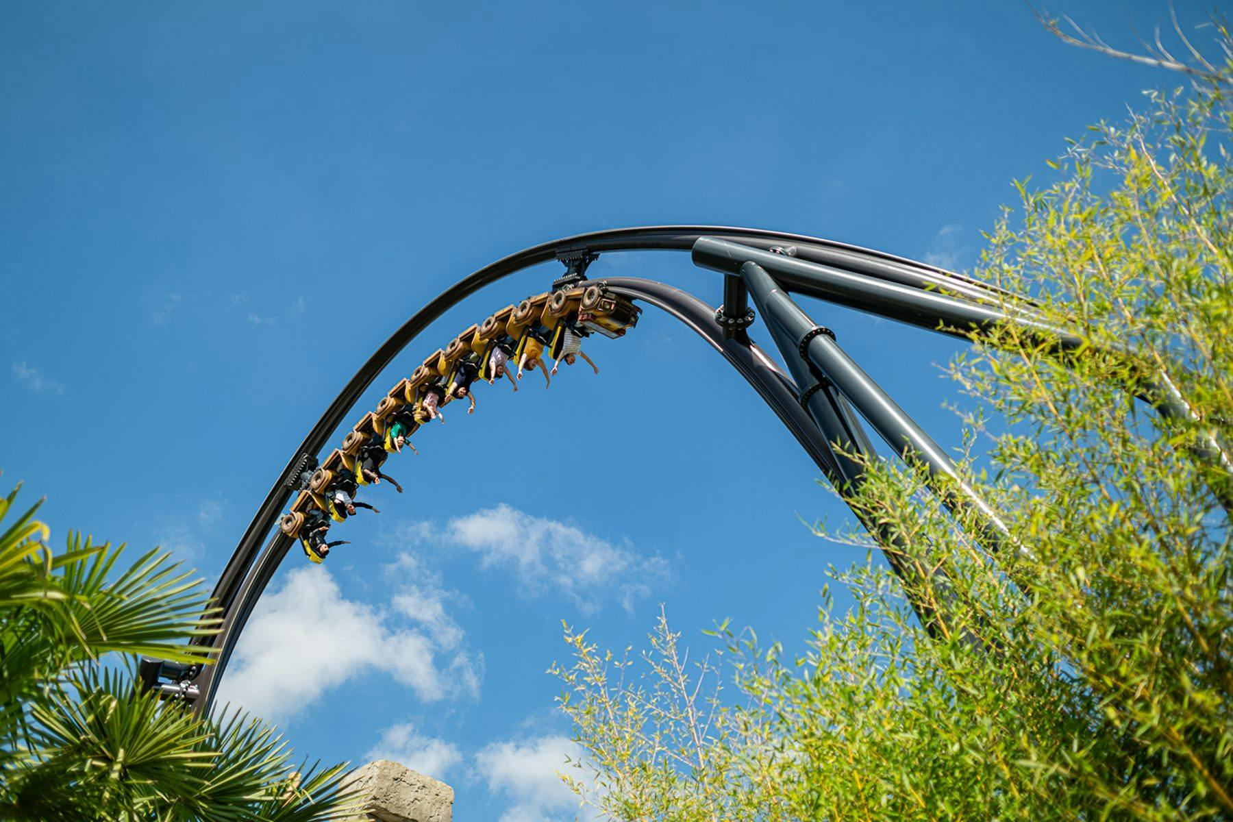 A rollercoaster with people on it upside-down in a loop, against a blue sky with clouds and some trees visible.