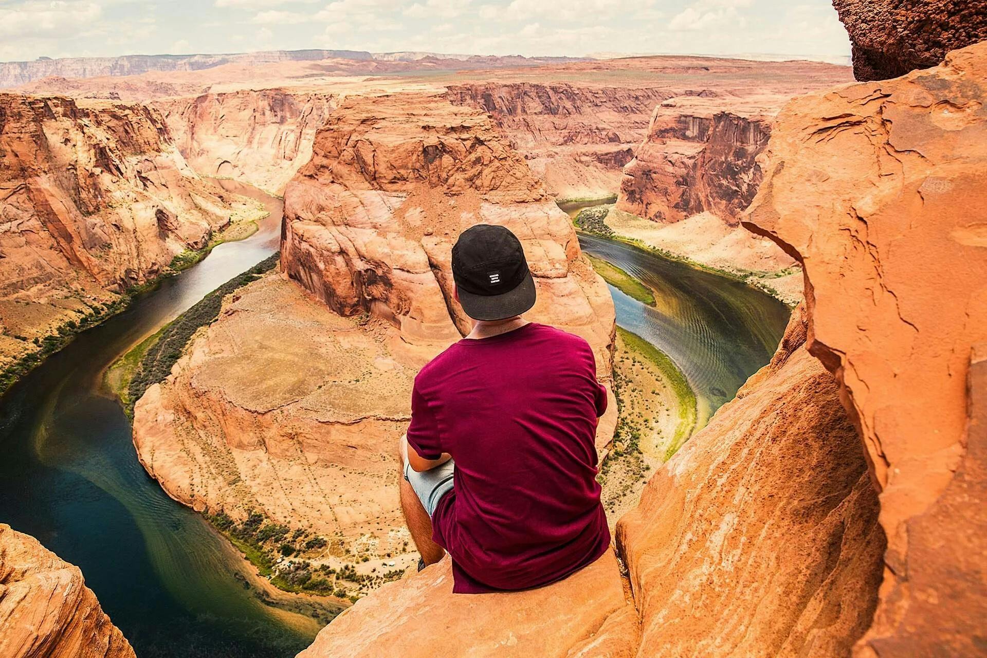 Person in a red shirt and black cap sits on a cliff overlooking a winding river and canyon landscape under a partly cloudy sky.