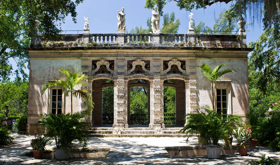 Historic stone building with arched doorways, statues on the roof, and decorative columns. Surrounded by lush greenery and palm trees.